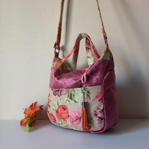 May include: A floral print shoulder bag with a matching strap and handles. The bag features a combination of pink, peach, and green floral patterns on a beige background, with a solid pink top and sides. An orange tassel hangs from the zipper.