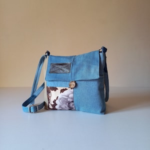 Könnte beinhalten: Eine blaue Jeans-Umhängetasche mit einem floralen Stoff-Patch und einem Holz-Knopf. Die Tasche hat einen Klappenverschluss und einen langen verstellbaren Riemen.