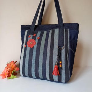 Op de afbeelding: Denim tote bag met verticale strepen in blauw- en grijstinten. Een rode bloemenapplicatie en kleine bloemblaadjes sieren de voorkant. De tas heeft lange hengsels en een decoratieve kwast met kralen.