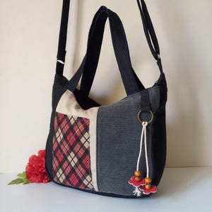 Puede incluir: Bolso de hombro patchwork con correa y asa negras. El bolso presenta un estampado de cuadros en rojo, beige y negro, con un panel de mezclilla y beige. Una borla decorativa con cuentas de madera y detalles de flores rojas cuelga de un anillo de metal.