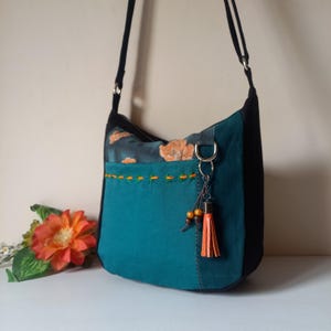 May include: A teal and black shoulder bag with a floral pattern on the top. The bag has a decorative orange tassel and a stitched pocket. The strap is black.