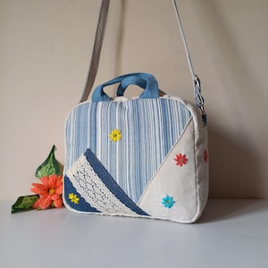 Puede incluir: Bolso de hombro cuadrado hecho a mano con un patrón de rayas azules y blancas. El bolso presenta detalles de mezclilla, ribete de encaje y flores bordadas en amarillo, rojo y azul. Tiene un asa de mezclilla y una correa ajustable.