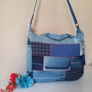 Puede incluir: Bolso de hombro de mezclilla patchwork con una correa larga. El bolso presenta varios tonos de parches de mezclilla azul, incluyendo telas a cuadros y lisas. Una pequeña borla azul y un llavero cuelgan del lateral. El bolso mide unos 30 cm de ancho y 25 cm de alto.