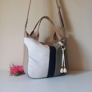 May include: A beige and white tote bag with a black stripe and a decorative tassel. The bag has a shoulder strap and two handles. The bag is made of canvas material and has a floral pattern on the side.