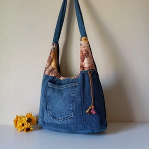以下が含まれることがあります： ブラウン、ベージュ、イエローの花柄生地をあしらったショルダーストラップと大きな外ポケットが特徴のブルーデニムのホーボーバッグ。バッグの側面には、装飾的なビーズが付いた茶色のコードがぶら下がっています。