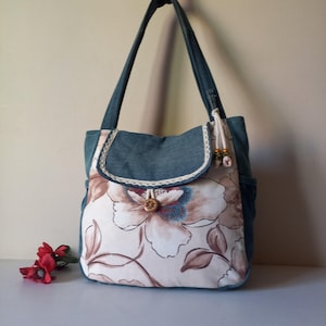 Könnte beinhalten: Eine Schultertasche aus blauem Jeansstoff kombiniert mit einem cremefarbenen Einsatz, der einen großen braun-blauen Blumendruck zeigt. Die Tasche verfügt über einen Klappverschluss mit Holzknopf, dekorative hängende Perlen und zwei Schulterriemen.
