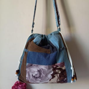 Può includere: Borsa a tracolla patchwork con chiusura a coulisse. La borsa presenta pannelli in denim, velluto a coste marrone e tessuto floreale. Coulisse bianche con perline di legno e un cinturino in denim completano il design. La borsa è larga circa 30 cm e alta 36 cm.