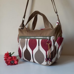 May include: A brown and patterned shoulder bag with a zipper closure and a decorative key ring. The bag features a unique pattern of red, white, and brown shapes. It has a brown handle and a long adjustable strap.