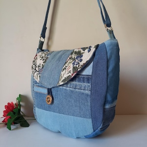 May include: A denim crossbody bag with a patchwork design. The bag features various shades of blue denim, with a flap adorned with floral fabric. A decorative button secures the flap, and the bag has a long, adjustable strap.
