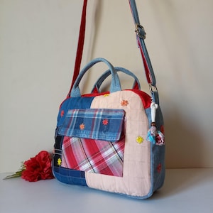Puede incluir: Bolso de hombro patchwork con telas vaqueras, rojas y a cuadros. El bolso tiene un bolsillo delantero, asas superiores y una correa ajustable. Flores bordadas adornan el bolso, añadiendo un toque decorativo.