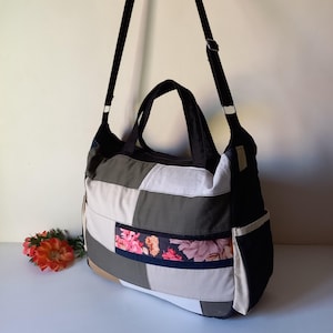 May include: A patchwork tote bag with a black shoulder strap and handles. The bag features a mix of neutral colors, including white, gray, and beige, with a floral accent. The bag has a side pocket and is designed for carrying items.