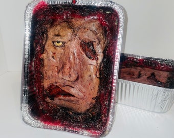 May include: A gruesome Halloween prop featuring a realistic, disfigured face in a rectangular aluminum foil pan. The face is a mix of flesh tones with a single visible yellow eye and a bloody, torn appearance. A smaller pan with similar contents is next to it.