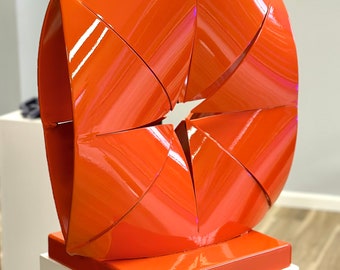 May include: A vibrant orange abstract sculpture with a glossy finish. The artwork features a geometric design with multiple folded panels, mounted on a square base. The sculpture is displayed on a white pedestal.