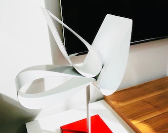 May include: A white metal sculpture of a stylized flower with a red square base. The sculpture is made of multiple curved pieces of metal that are joined together to form a flower shape.