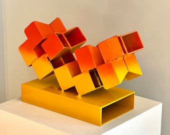 May include: Abstract sculpture made of metal cubes, painted in a gradient of orange and yellow. The sculpture is mounted on a yellow base.