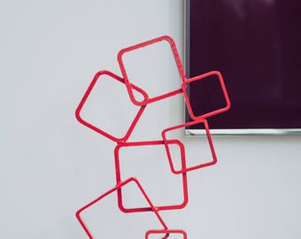 May include: A red metal sculpture made of stacked and overlapping squares. The sculpture is on a white surface and has a square base.