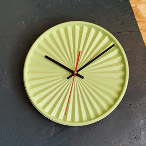 Puede incluir: Un reloj de pared redondo verde claro con un diseño en forma de abanico. Tiene manecillas negras para las horas y los minutos, y un segundero rojo. El reloj está montado en una pared texturizada gris oscuro.
