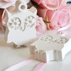 Może przedstawiać: Dwa białe ceramiczne ozdoby w kształcie sowy z detalicznymi oczami i różową wstążką. Sowy wiszą na wstążce.