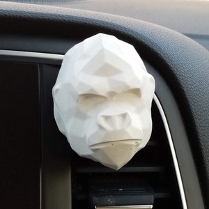 Può includere: Un diffusore per auto a forma di testa di gorilla, stampato in 3D, bianco e geometrico, appeso a una bocchetta di ventilazione dell'auto.