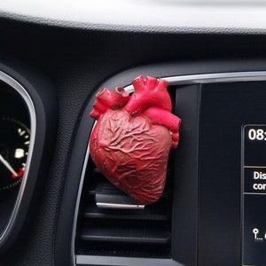 Puede incluir: Un ambientador de aire en forma de corazón anatómico rojo colgado del respiradero del tablero de un coche. El tablero del coche es negro y tiene una pantalla digital que muestra la hora 08:13 y el texto "Distância sem consumo".