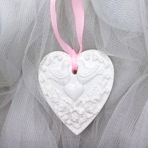 May include: White ceramic heart ornament with a raised design of two birds and a heart, hanging from a pink ribbon on a white tulle background.