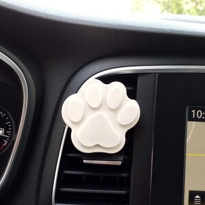 May include: White paw-shaped car air freshener hanging from a car's air vent. The air freshener is in the shape of a paw print with four toes and a pad. The air vent is black and has a silver trim. The car's dashboard is black.