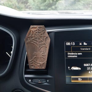 Puede incluir: Un clip de ventilación de coche en forma de ataúd, de color bronce. El clip presenta un diseño de araña y telaraña. El salpicadero del coche es negro y la pantalla muestra la hora: 08:13. El botón de arranque/parada del coche es visible.
