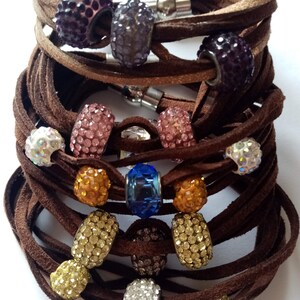 May include: A stack of brown leather bracelets with various colored crystal beads. The beads are round and have a sparkly finish. The bracelets are tied together with a silver clasp.