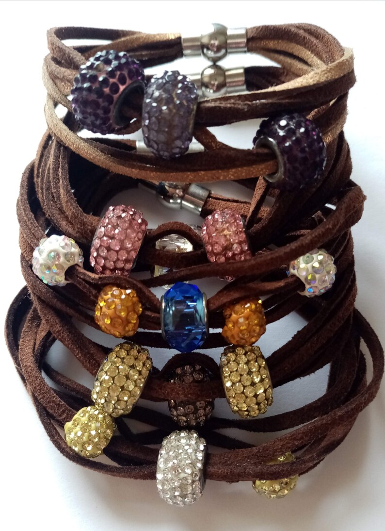 May include: A stack of brown leather bracelets with various colored crystal beads. The beads are round and have a sparkly finish. The bracelets are tied together with a silver clasp.