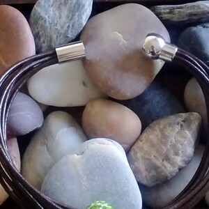 May include: A brown leather bracelet with silver magnetic clasps. The bracelet is made of multiple strands of leather and is shown on a background of smooth stones.