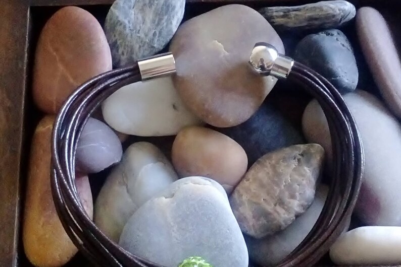 May include: A brown leather bracelet with silver magnetic clasps. The bracelet is made of multiple strands of leather and is shown on a background of smooth stones.