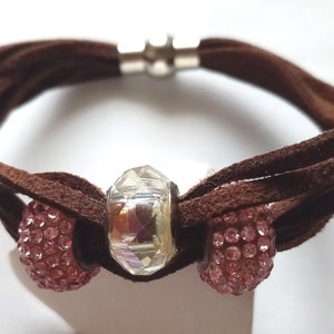 May include: A brown suede bracelet with a magnetic clasp and three beads. The center bead is clear and faceted. The two outer beads are pink and covered in small rhinestones.