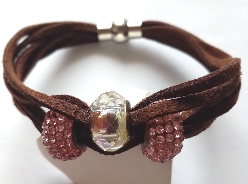 May include: A brown suede bracelet with a magnetic clasp and three beads. The center bead is clear and faceted. The two outer beads are pink and covered in small rhinestones.