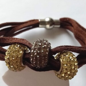 May include: A brown suede bracelet with a magnetic clasp and three beaded charms. The charms are round and covered in clear rhinestones. Two of the charms are gold and one is silver.