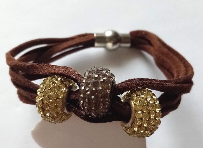 May include: A brown suede bracelet with a magnetic clasp and three beaded charms. The charms are round and covered in clear rhinestones. Two of the charms are gold and one is silver.