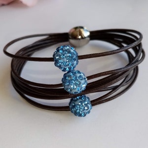 May include: A brown leather bracelet with three blue crystal beads. The bracelet is made of multiple strands of leather and has a magnetic clasp.