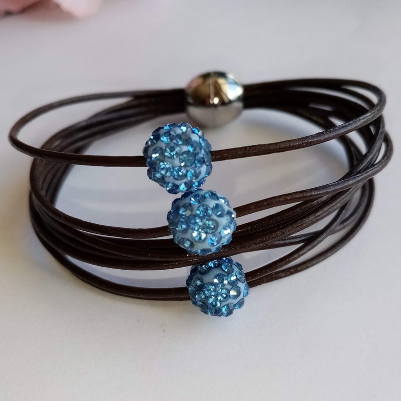May include: A brown leather bracelet with three blue crystal beads. The bracelet is made of multiple strands of leather and has a magnetic clasp.