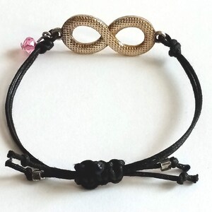 May include: A black cord bracelet with an infinity symbol charm. The charm is gold-colored and has a textured surface. A small pink crystal bead is attached to the cord.