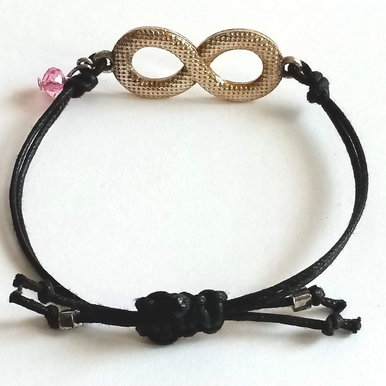 May include: A black cord bracelet with an infinity symbol charm. The charm is gold-colored and has a textured surface. A small pink crystal bead is attached to the cord.