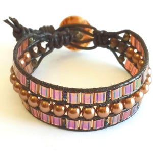 Könnte beinhalten: Ein braunes Lederarmband mit einer doppelten Reihe kupferfarbener Perlen und schimmernden rosa und violetten Glasfliesen.