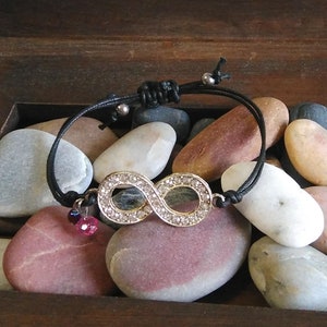 May include: A black string bracelet with a silver infinity charm set with clear rhinestones. The bracelet has two small pink and purple beads hanging from it.