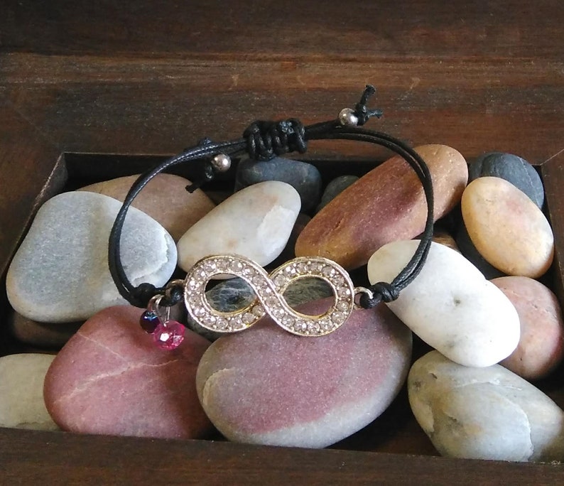 May include: A black string bracelet with a silver infinity charm set with clear rhinestones. The bracelet has two small pink and purple beads hanging from it.