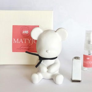 May include: White ceramic bear figurine with a black ribbon around its neck. The bear is sitting on a white surface. The background includes a white box with the text "MATYJO jewellery and pewter gifts" in pink and black.