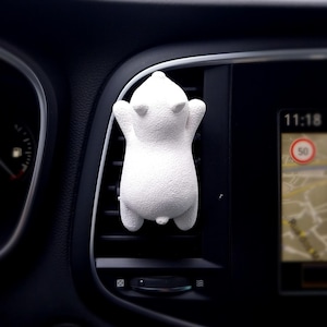 Può includere: Un deodorante per auto a forma di orso bianco è attaccato a una presa d'aria dell'auto. Il deodorante ha una superficie strutturata ed è posizionato davanti a un cruscotto nero con un display digitale che mostra l'ora e una mappa.