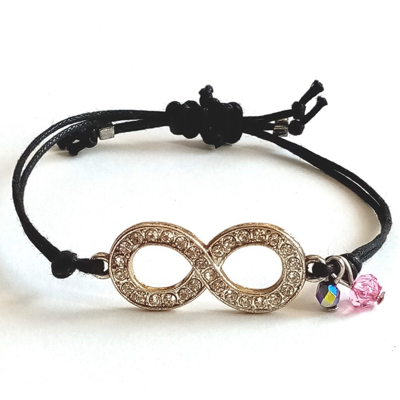 May include: Black string bracelet with an infinity symbol charm. The charm is gold-toned and encrusted with clear rhinestones. The bracelet also features a pink and iridescent crystal charm.
