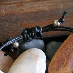 May include: Close-up of a black cord bracelet with silver beads. The bracelet is tied in a knot and is resting on a pile of smooth, gray and brown stones.