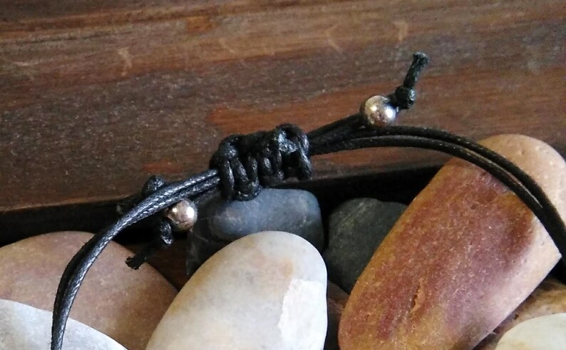 May include: Close-up of a black cord bracelet with silver beads. The bracelet is tied in a knot and is resting on a pile of smooth, gray and brown stones.