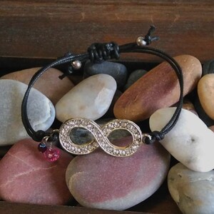 May include: A black cord bracelet with an infinity symbol charm. The charm is silver and has clear rhinestones. The bracelet is accented with two small pink and purple beads.