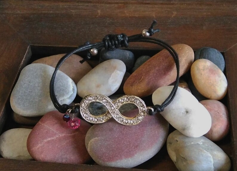 May include: A black cord bracelet with an infinity symbol charm. The charm is silver and has clear rhinestones. The bracelet is accented with two small pink and purple beads.