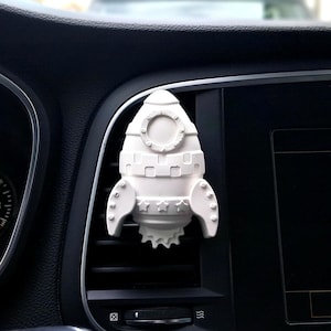 May include: White ceramic rocket-shaped car air freshener with a star design. The air freshener is attached to a car's air vent.